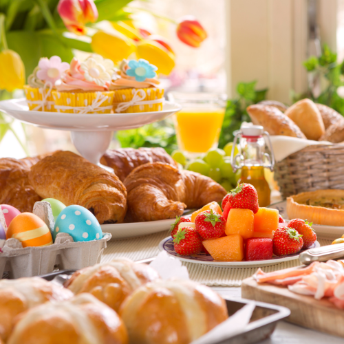 The image features a festive breakfast spread with croissants, colorful decorated eggs, fruit, pastries, juice, and flowers in the background.