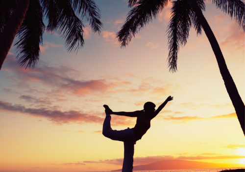 A person is doing a yoga pose on the beach at sunset, framed by palm trees, with a vibrant sky in the background.