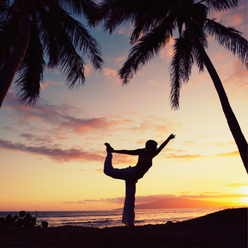 A person is doing a yoga pose on the beach at sunset, framed by palm trees, with a vibrant sky in the background.