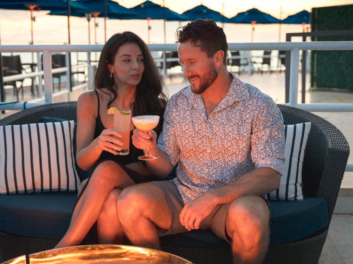 A couple sits on an outdoor sofa, enjoying cocktails and each other's company, with a sunset view and patio umbrellas in the background.
