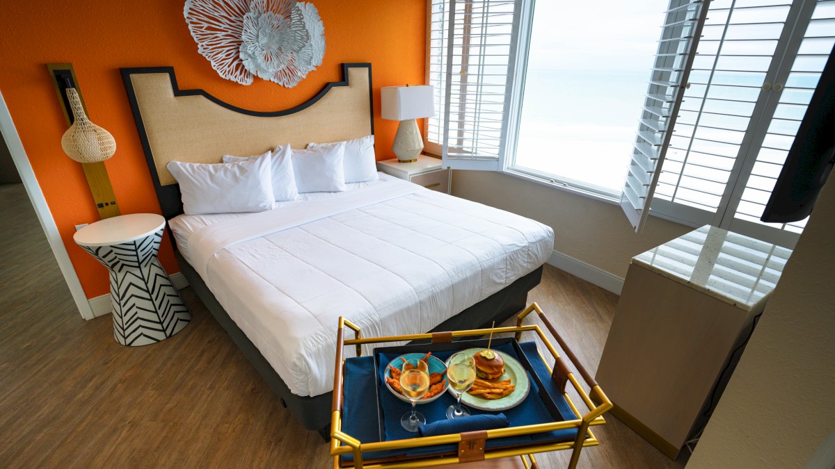 A modern bedroom features a large bed with white linens, colorful decor, and a tray with food and drinks by the window with blinds.