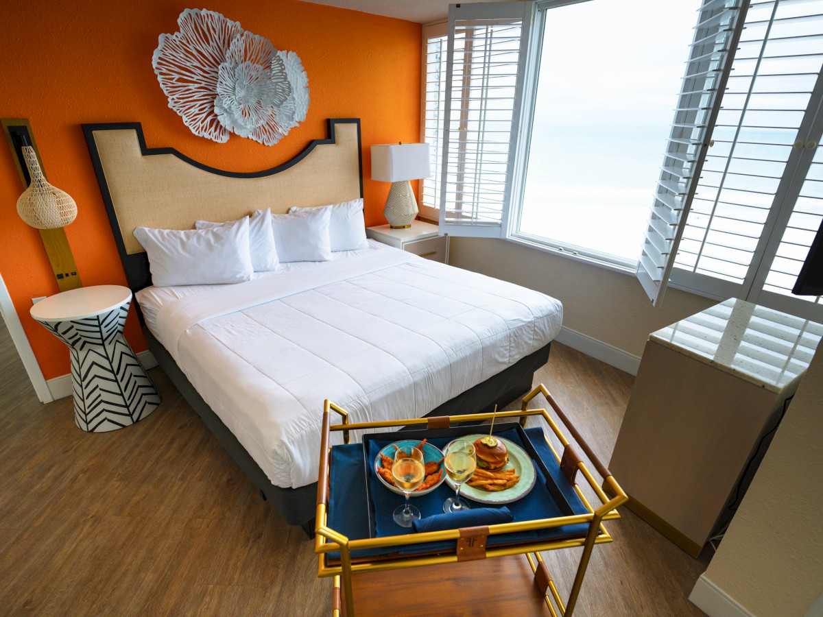 A modern bedroom features a large bed with white linens, colorful decor, and a tray with food and drinks by the window with blinds.
