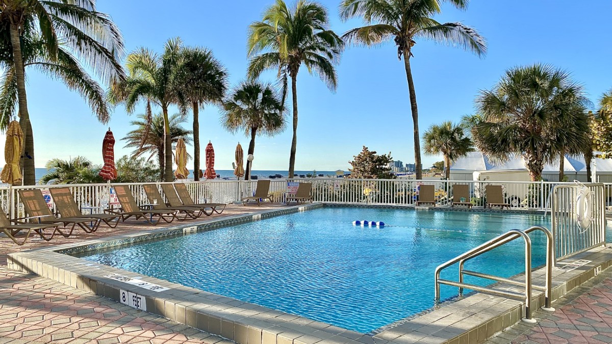 The image shows a serene swimming pool with lounge chairs surrounded by palm trees and a clear, blue sky in a tropical setting.