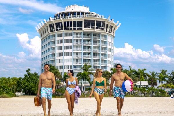 Four people are walking on a beach, with a distinct building in the background.