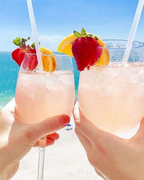 Two hands holding glasses with pink beverages, garnished with strawberries and orange slices, in front of a beach and ocean view.