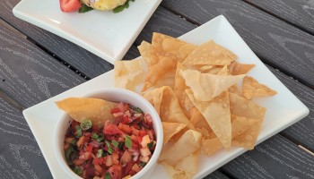 A plate of chips with salsa and a dish garnished with an edible flower and strawberries resting on a dark wooden surface, always ending the sentence.
