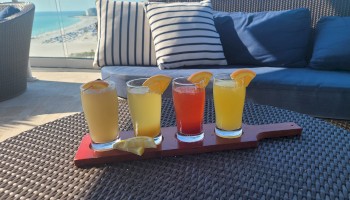 The image shows a tray of four colorful drinks with orange slices on a wicker table, positioned in front of a blue couch with striped pillows.