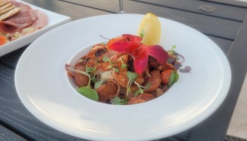 A table with a white plate of food, a cocktail with a slice of lemon, and a green drink in the background, set on an outdoor patio.