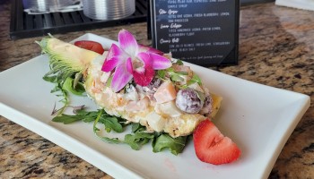 The image shows a plate with a beautifully presented dish garnished with flowers and strawberries, placed on a countertop beside a drink menu.