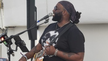 A musician is performing with a guitar and microphone. A neon sign in the background reads, 