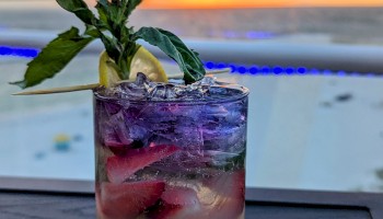 A cocktail garnished with basil and a lemon slice sits on a table by a pool, with a serene sunset in the background.