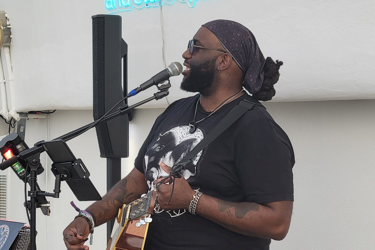 A musician wearing a bandana and sunglasses is playing a guitar and singing on stage. A neon sign reads 