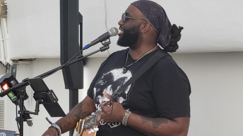 A musician wearing a bandana and sunglasses is playing a guitar and singing on stage. A neon sign reads 