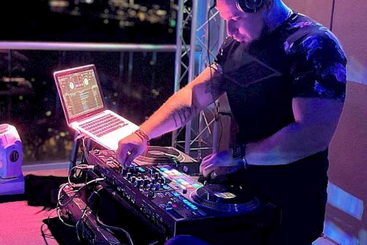 A person is DJing at a booth with a laptop and other equipment, wearing headphones. The scene appears to be in a dimly lit setting.