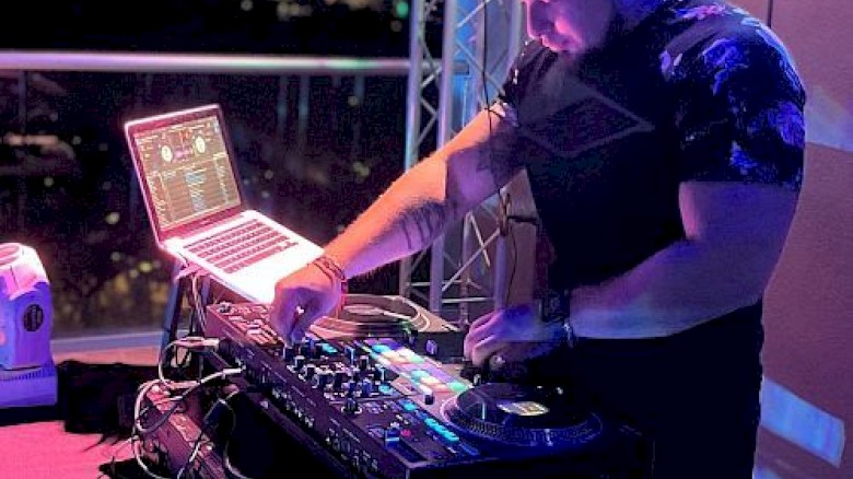 A person is DJing at a booth with a laptop and other equipment, wearing headphones. The scene appears to be in a dimly lit setting.