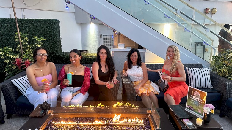 Five women are sitting around a fire pit, holding drinks and smiling, in an outdoor seating area with greenery and a staircase.