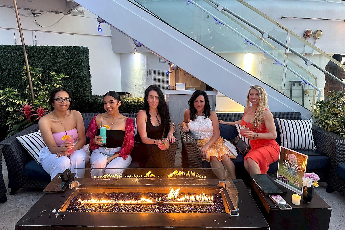 Five women are sitting around a fire pit, holding drinks and smiling, in an outdoor seating area with greenery and a staircase.