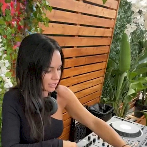 A woman is using DJ equipment in an outdoor setting, surrounded by greenery and wooden paneling.