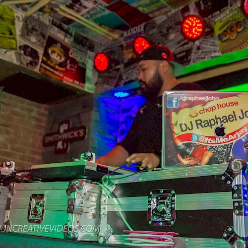 A DJ is performing in a colorful, poster-filled room with equipment and a visible sign reading 