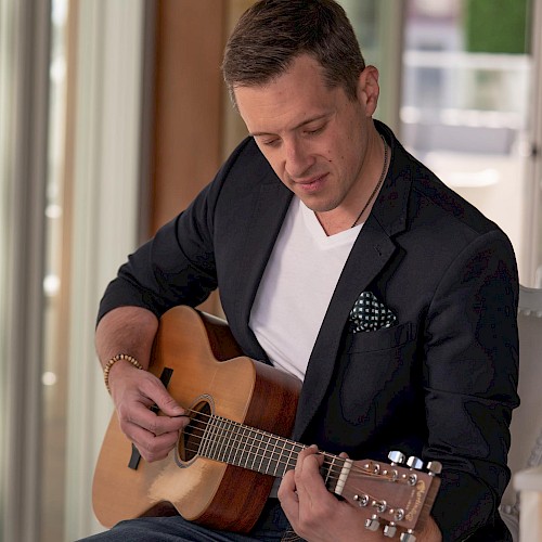 A person is sitting and playing an acoustic guitar, wearing a dark blazer and a white shirt, focused on the instrument.