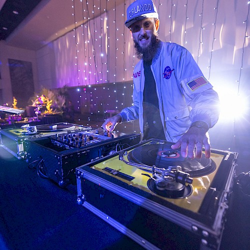 A DJ is performing on a set of turntables in a dimly lit room with string lights in the background and a relaxed atmosphere.