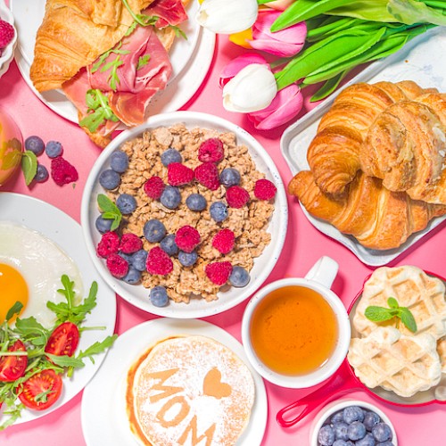 The image features a breakfast spread with croissants, a fried egg, cereal with berries, pancakes, waffles, coffee, juice, and a floral bouquet.