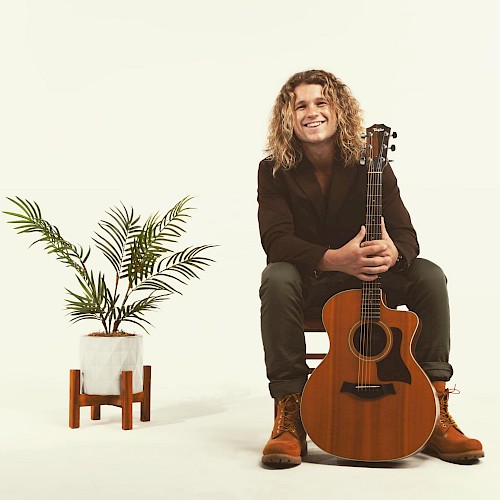 A person is seated with a guitar, smiling at the camera. A potted plant is on their left, both set against a plain white background.