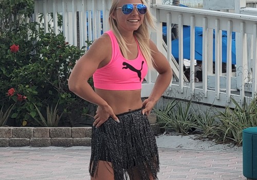 A person wearing a pink sports top and a fringe skirt is standing outside near a white fence with tropical plants and a beach setting.