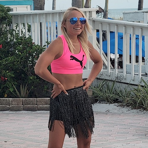 A person wearing a pink sports top and a fringe skirt is standing outside near a white fence with tropical plants and a beach setting.