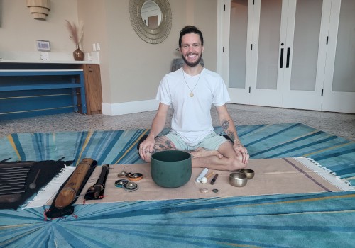 A person sits cross-legged on a mat with various sound healing instruments, such as singing bowls and chimes, laid out in front of them.