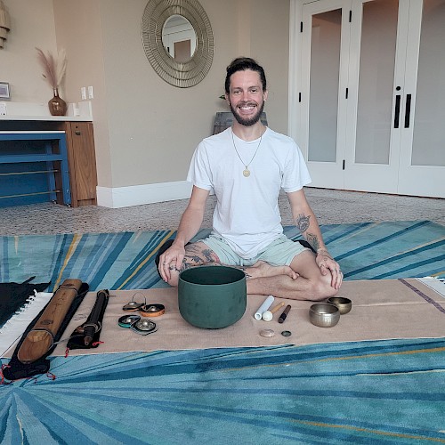 A person sits cross-legged on a mat with various sound healing instruments, such as singing bowls and chimes, laid out in front of them.