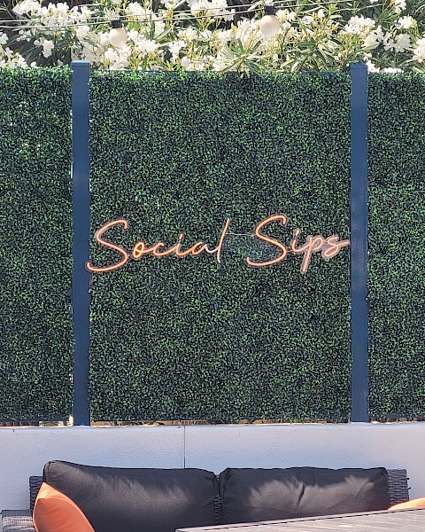 The image shows a seating area in an outdoor setting with a green hedge wall and a neon sign that says 