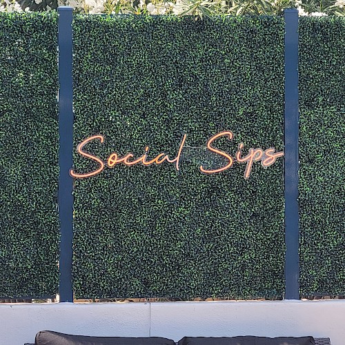 The image shows a seating area in an outdoor setting with a green hedge wall and a neon sign that says 