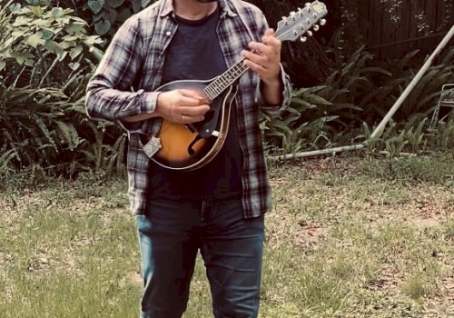 A man is standing in a grassy backyard, playing a string instrument, possibly a mandolin. He wears a plaid shirt and jeans, against a backdrop of greenery.