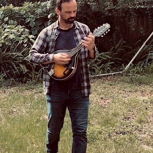 A man is standing in a grassy backyard, playing a string instrument, possibly a mandolin. He wears a plaid shirt and jeans, against a backdrop of greenery.