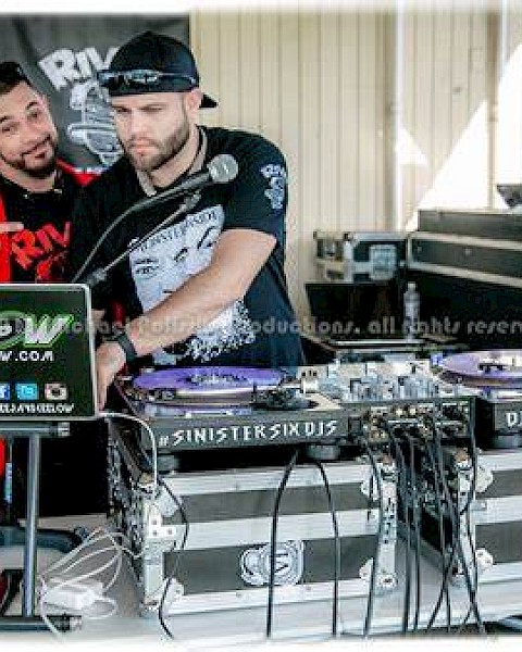 Two DJs are working with turntables and a laptop at an event. One is pointing at the other’s laptop while the other adjusts the equipment.