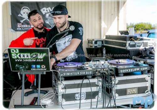 Two DJs are working with turntables and a laptop at an event. One is pointing at the other’s laptop while the other adjusts the equipment.