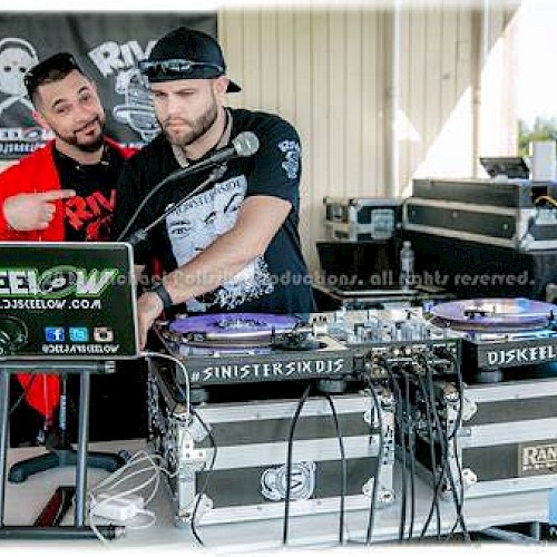 Two DJs are working with turntables and a laptop at an event. One is pointing at the other’s laptop while the other adjusts the equipment.