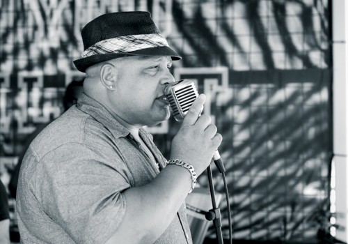 A person wearing a hat and a short-sleeved shirt is singing into a retro-style microphone, with an abstract background behind them.