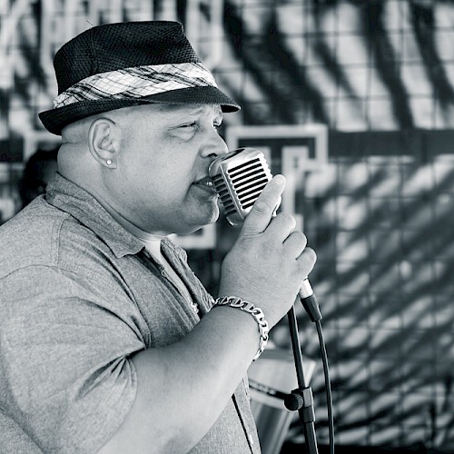 A person wearing a hat and a short-sleeved shirt is singing into a retro-style microphone, with an abstract background behind them.