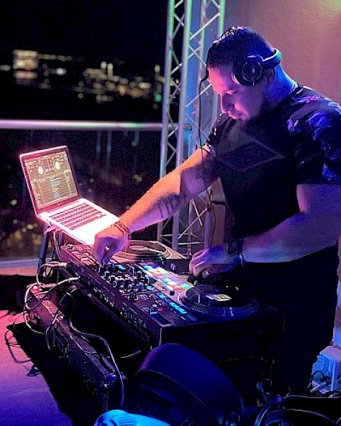 A DJ is at work at a mixing console with a laptop, wearing headphones. The setting appears to be a nighttime event with ambient lighting.
