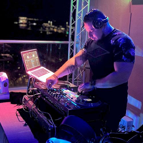 A DJ is at work at a mixing console with a laptop, wearing headphones. The setting appears to be a nighttime event with ambient lighting.