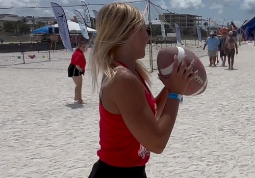 A lady is holding a football, while others are playing on a sandy beach, with buildings and event banners in the background.