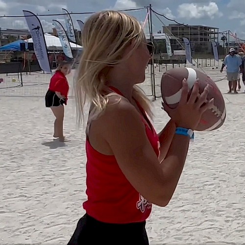 A lady is holding a football, while others are playing on a sandy beach, with buildings and event banners in the background.