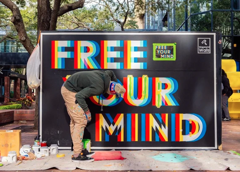 A man is painting a colorful sign on a black background that reads "FREE YOUR MIND" in a public place.