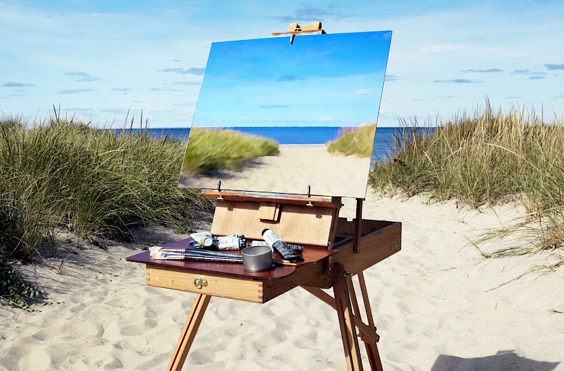 An easel with a painting of a sandy beach and ocean waves stands on the beach, surrounded by dunes under a clear blue sky.