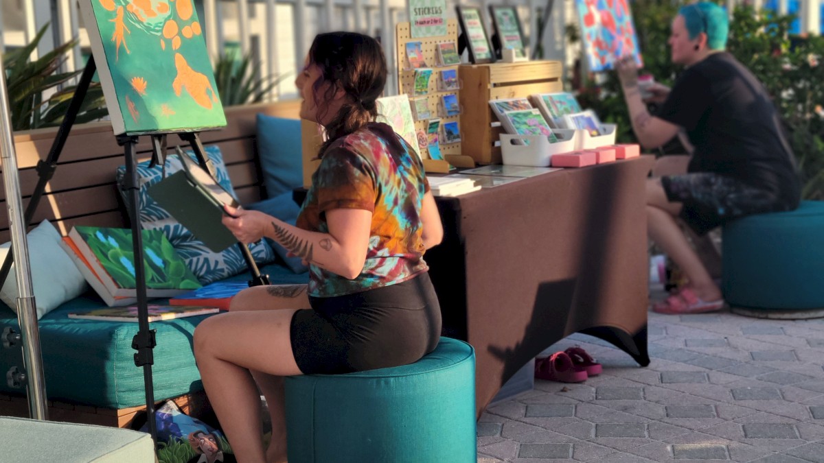 Two people are painting at an outdoor setup with easels and various art supplies. They are seated on stools under umbrellas, near a fence and palm trees.