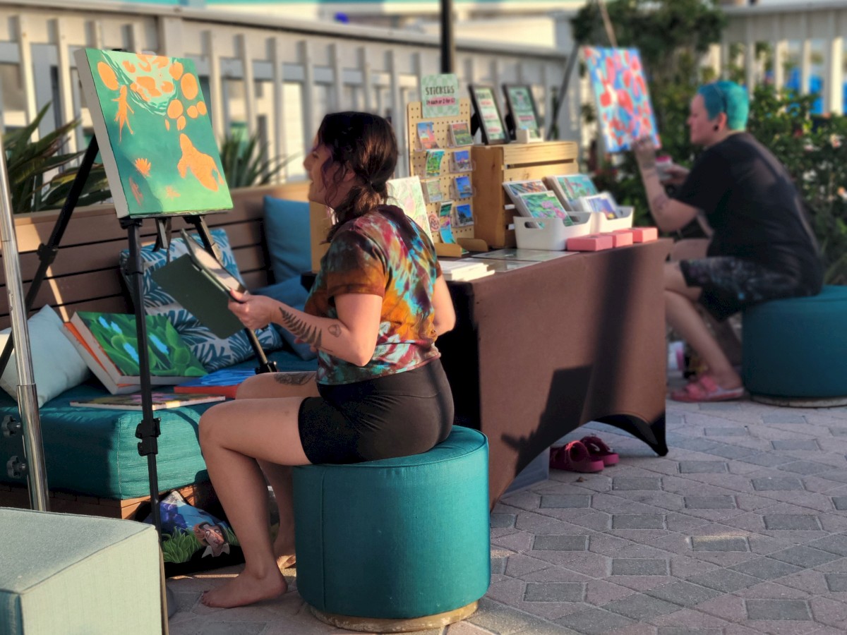 Two people are painting at an outdoor setup with easels and various art supplies. They are seated on stools under umbrellas, near a fence and palm trees.