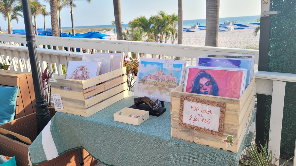 The image shows a beachside table with art prints in wooden crates, a cash box, and a sign reading 