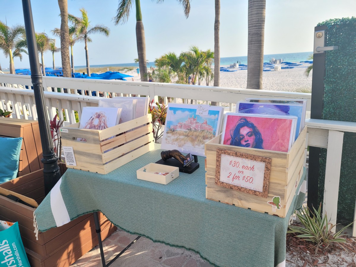 The image shows a beachside table with art prints in wooden crates, a cash box, and a sign reading 